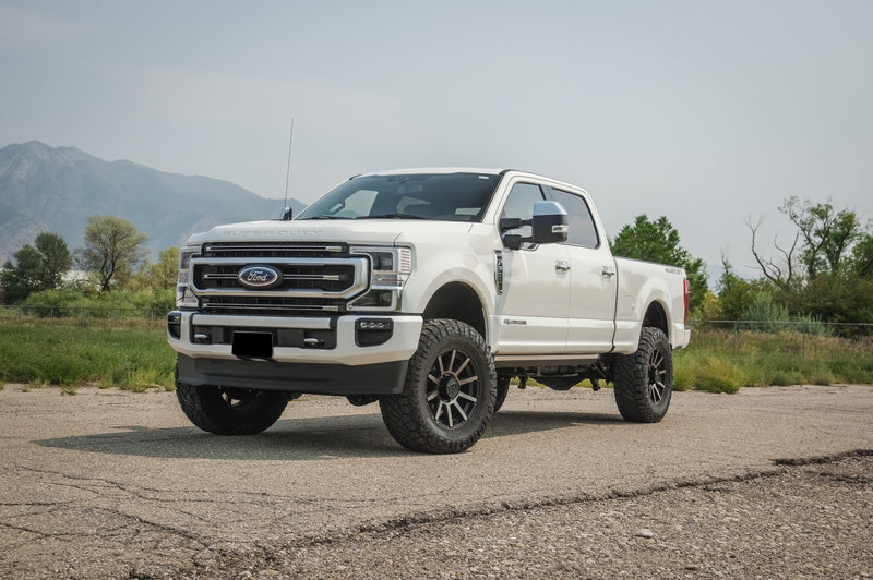 ReadyLIFT Coil Spring Suspension System installed on a Ford F-250 Super Duty: white Ford Super Duty crew cab lifted on black wheels on a paved lot with mountains behind