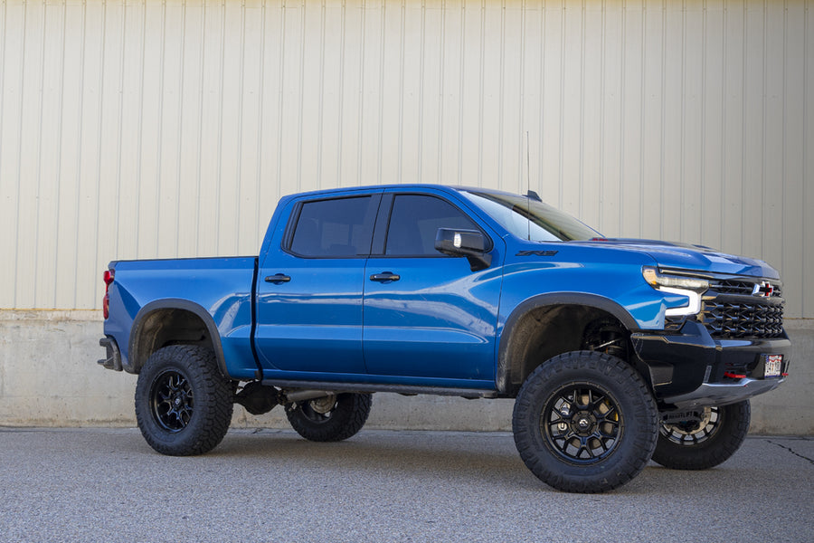 ReadyLIFT Lift Kit installed on a Chevrolet Silverado 1500 ZR2: blue Chevrolet Silverado ZR2 crew cab lifted on black wheels and all-terrain tires beside a concrete wall