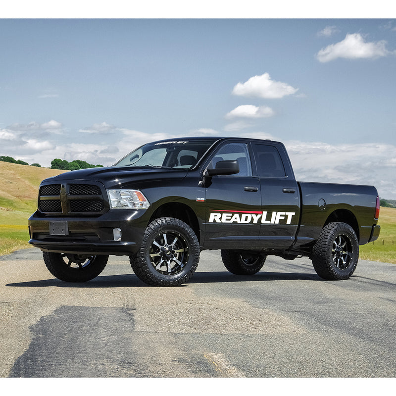 ReadyLIFT SST Lift Kit installed on a Ram 1500: A black lifted Ram 1500 Quad Cab with a ReadyLIFT door decal parked on a paved road under blue sky