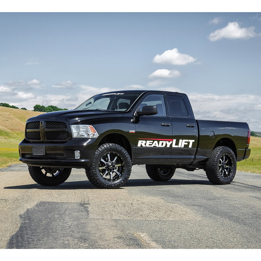 ReadyLIFT SST Lift Kit installed on a Ram 1500: A black lifted Ram 1500 Quad Cab with a ReadyLIFT door decal parked on a paved road under blue sky