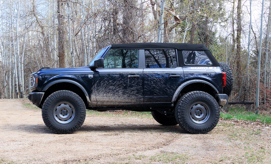ReadyLIFT SST Lift Kit installed on a Ford Bronco: A dark blue lifted Ford Bronco four-door with a soft top parked on gravel in front of trees, side view