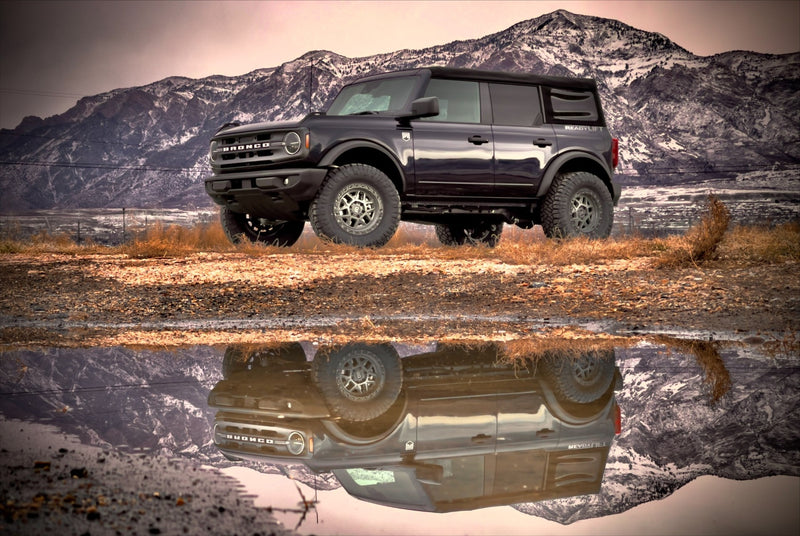 ReadyLIFT SST Lift Kit installed on a Ford Bronco: A dark gray lifted Ford Bronco four-door reflected in a puddle with mountains behind