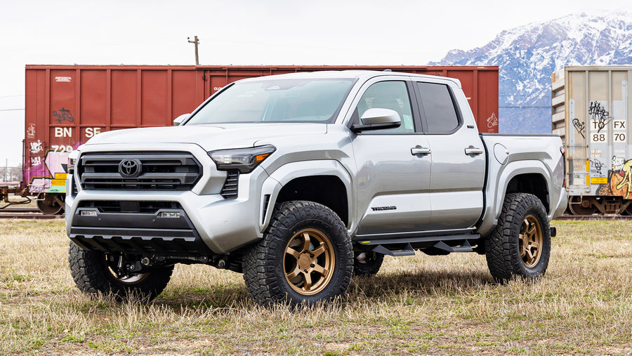 ReadyLIFT SST Lift Kit installed on a Toyota Tacoma: A silver lifted Toyota Tacoma Double Cab on bronze wheels parked on grass in front of rail cars