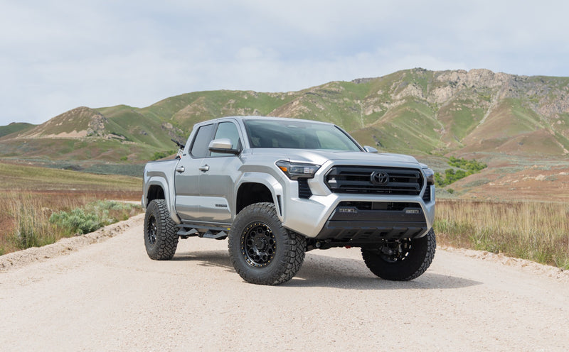 ReadyLIFT SST Lift Kit installed on a Toyota Tacoma: A silver lifted Toyota Tacoma Double Cab on a gravel road with green hills behind