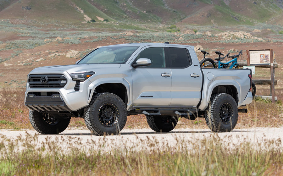 ReadyLIFT SST Lift Kit installed on a Toyota Tacoma: A silver lifted Toyota Tacoma Double Cab with bikes in the bed parked on a gravel road beside a sign