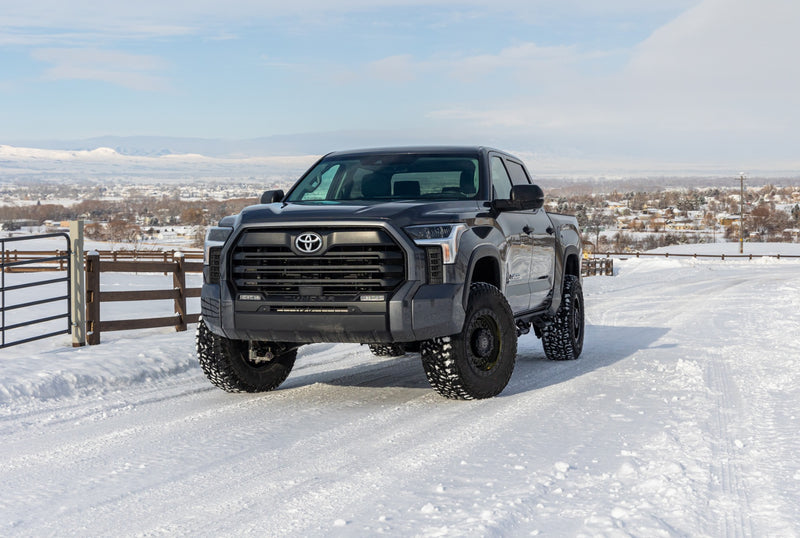 ReadyLIFT SST2.1 Suspension System installed on a Toyota Tundra: A gray lifted Toyota Tundra CrewMax parked on a snowy road beside a wooden fence