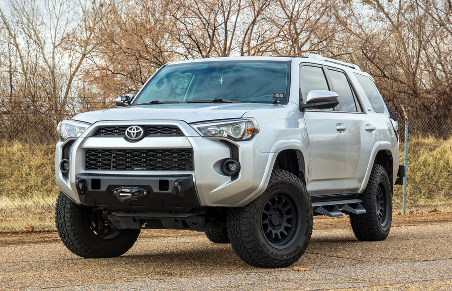 ReadyLIFT SST2.1 Suspension System installed on a Toyota 4Runner: A silver lifted Toyota 4Runner with a winch bumper parked on pavement in front of dry brush