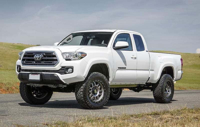 ReadyLIFT SST2.1 Suspension System installed on a Toyota Tacoma: A white lifted Toyota Tacoma Access Cab parked on a road beside a green field