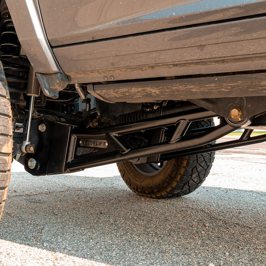 Big Lift Kits by ReadyLIFT: Black ReadyLIFT tubular radius arm installed under a pickup behind the front tire, seen from low on gravel.