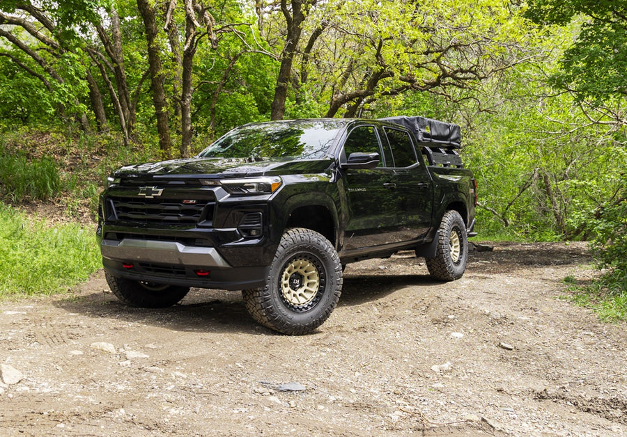 SST Performance Lift Kits by ReadyLIFT: A black lifted Chevrolet Colorado with bronze wheels parked on a dirt trail in green woods.