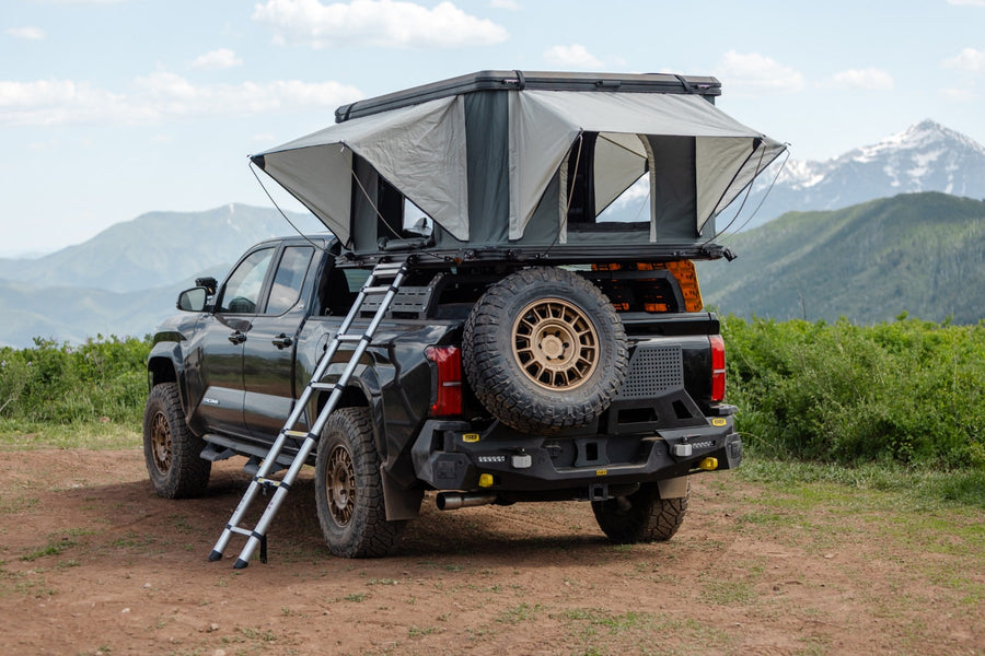 SST Performance Lift Kits by ReadyLIFT: A black lifted Toyota Tacoma with a bed rack, rooftop tent and ladder parked on dirt, rear view.