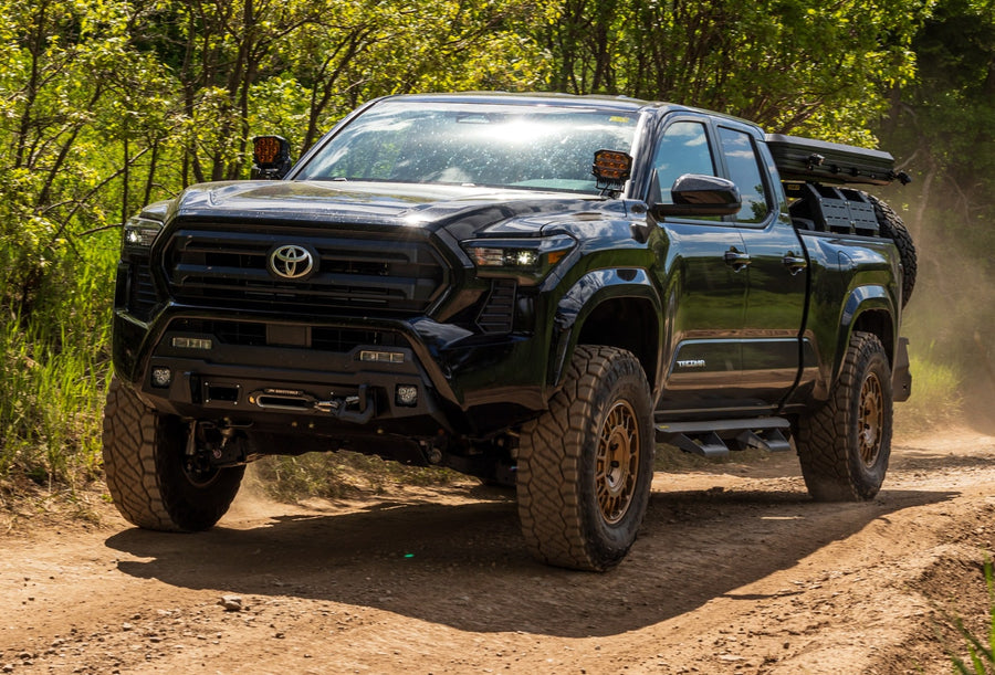 SST Performance Lift Kits by ReadyLIFT: A black lifted Toyota Tacoma with a bed rack driving on a dirt trail through trees.