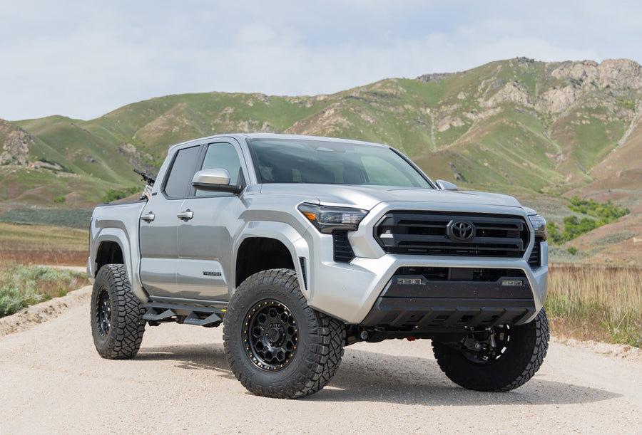 SST Performance Lift Kits by ReadyLIFT: A silver lifted Toyota Tacoma Double Cab on a gravel road with green hills behind.