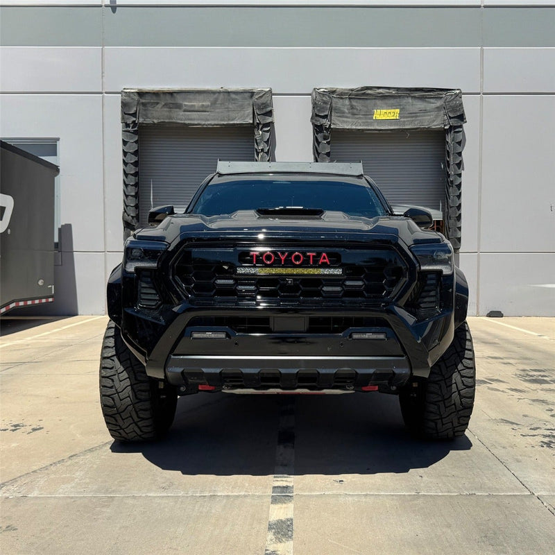 Rigid 20" SR-Series Amber Grille Kit installed on a Toyota Tacoma: front view of a black lifted Toyota Tacoma with red TOYOTA grille lettering and a 20" light bar in the grille, parked by loading docks
