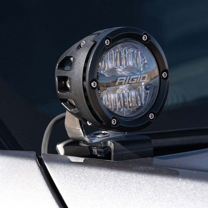 Close-up of the Rigid 360-Series 4" Drive A-Pillar Light Kit: close-up of a black 4" 360-Series pod with a clear lens on an A-pillar mount over a silver hood