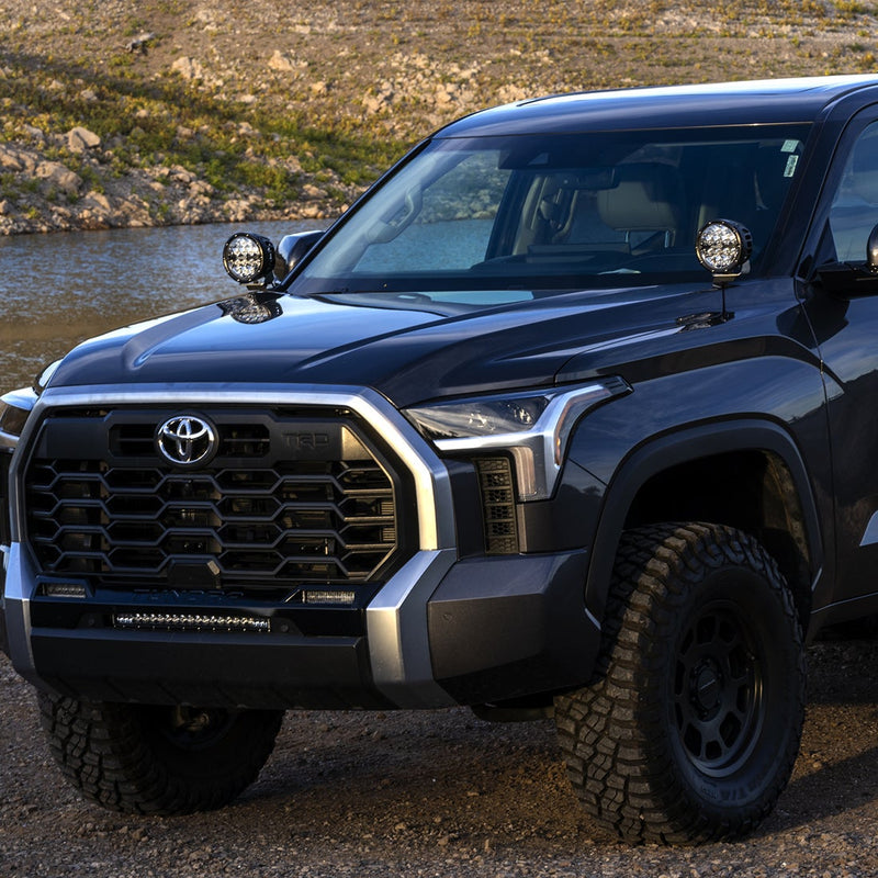 Rigid 360-Series 6" Drive A-Pillar Light Kit installed on a Toyota Tundra: front three-quarter view of a dark Toyota Tundra with a round LED light mounted at each A-pillar beside a lake