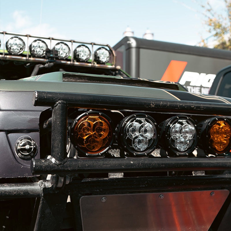 Rigid 360-Series Laser Optic 6" Pair installed on a vehicle: amber and clear 6" Rigid 360-Series lights mounted in a tube bumper of a green truck with more pods on its roof rack