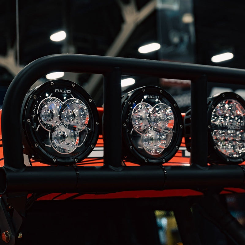 Rigid 360-Series Laser Optic 6" Pair installed on a vehicle: several 6" Rigid 360-Series lights with three-optic faces mounted on a black tube bar of a red vehicle indoors
