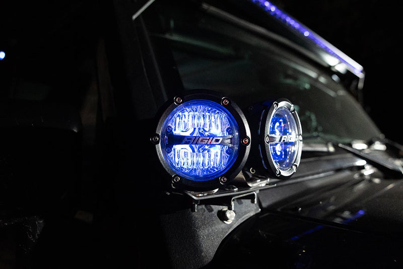 Rigid 360-Series PRO 4" Pair installed on a vehicle: two round LED pods mounted at the A-pillar of a dark vehicle glow with blue backlighting at night