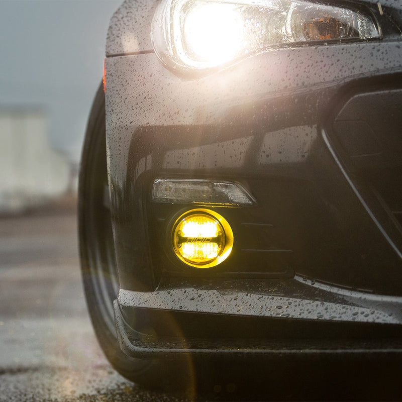 Rigid 360-Series PRO 4" SAE Pair installed on a Subaru BRZ: A round fog light glows yellow in the lower bumper of a dark, rain-beaded sports car