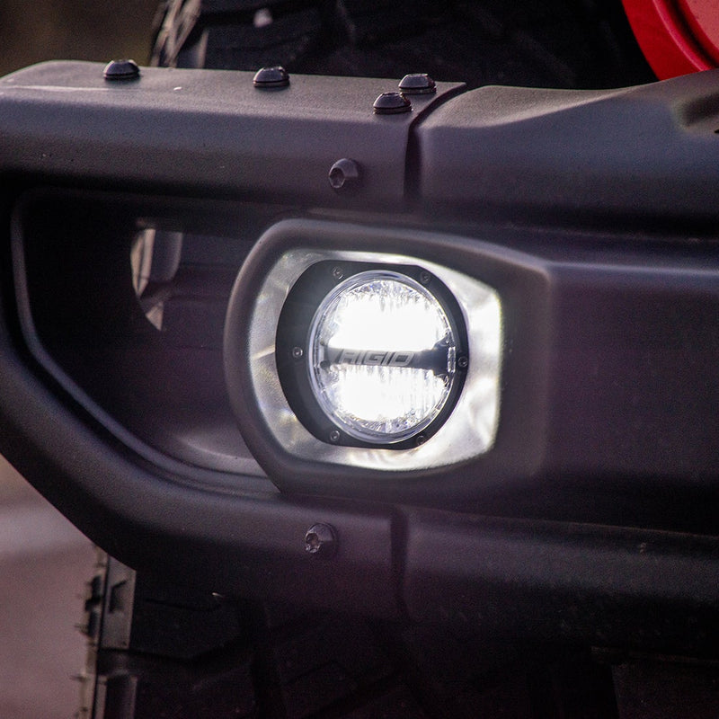 Rigid 360-Series PRO 4" SAE Pair installed on a vehicle: A round LED fog light glows white inside the fog pocket of a black bumper