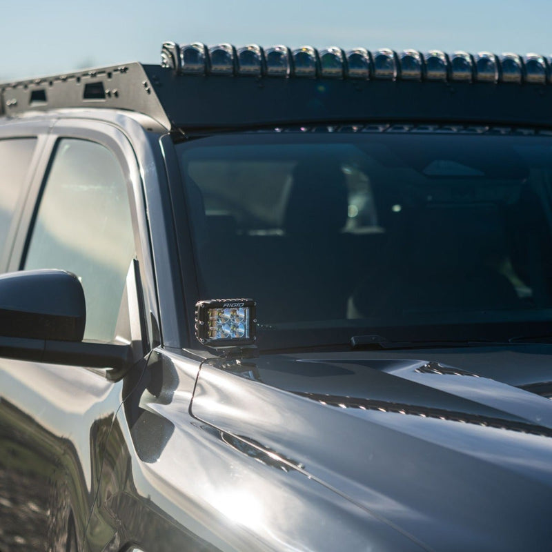 Rigid A-Pillar Bracket installed on a vehicle: close-up of a black cube LED light mounted at the A-pillar of a gray pickup below a roof rack light bar