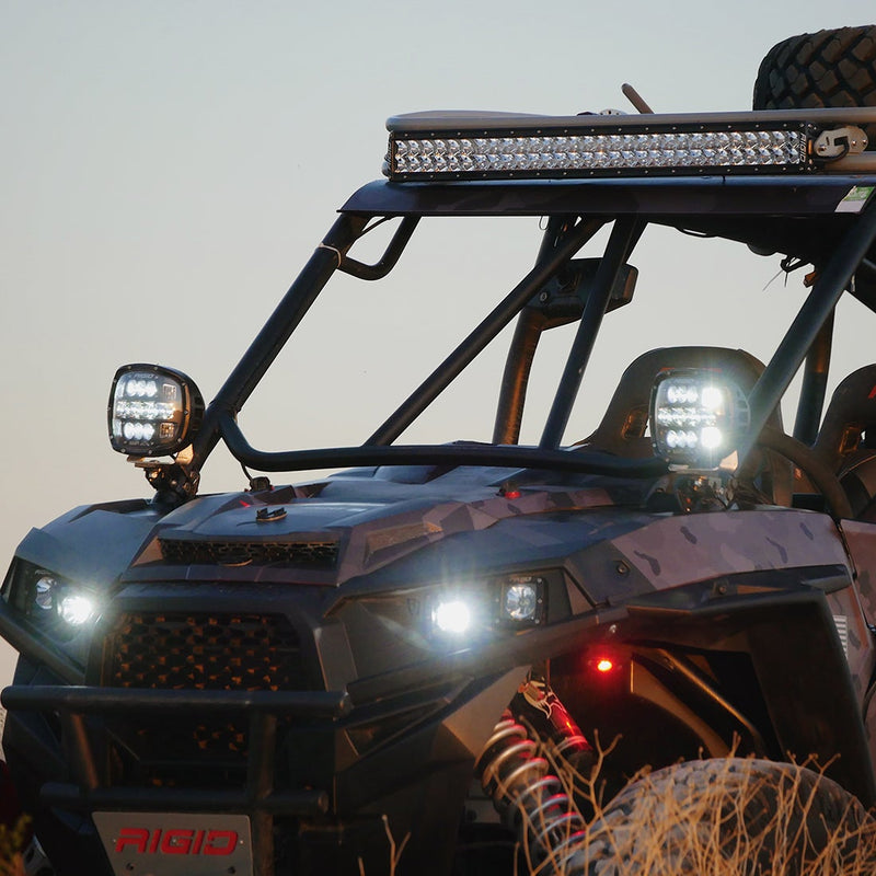Rigid Adapt XP Pair installed on a Polaris RZR: close front corner view of a side-by-side UTV with two white Adapt XP lights glowing on the A-pillars below a roof light bar