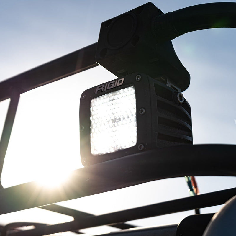 Close-up of the Rigid D-Series PRO Pods Pair: A D-Series PRO pod with a diffused lens shining white, mounted under a vehicle cage tube