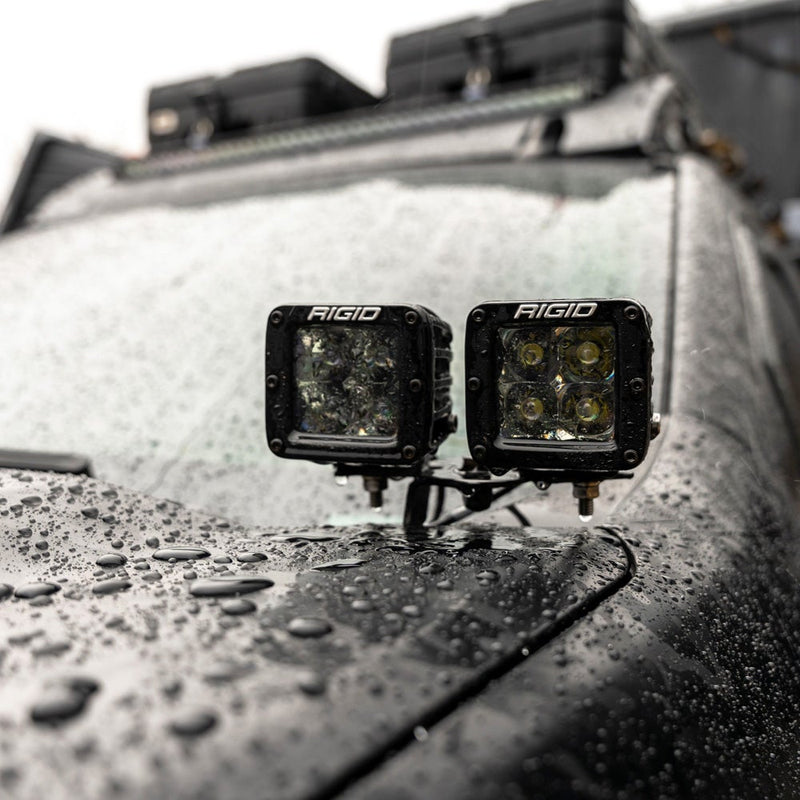Rigid D-Series PRO Pods Pair installed on a vehicle: A pair of D-Series PRO pods on a bracket at the base of a wet, dark vehicle windshield