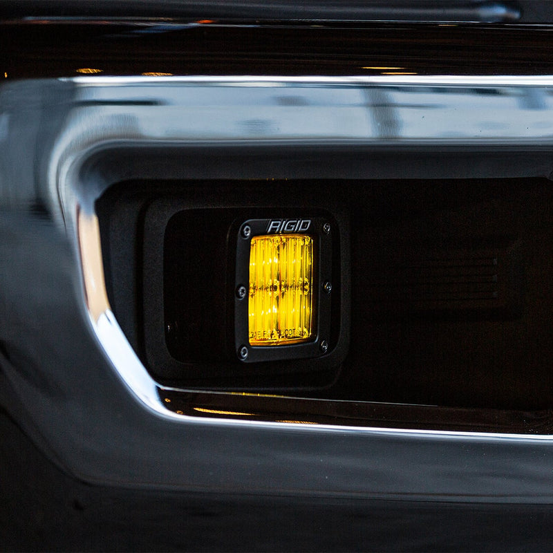 Rigid D-Series PRO SAE Pods Pair installed on a vehicle: A close view of an amber SAE pod glowing inside a black bumper fog pocket at night