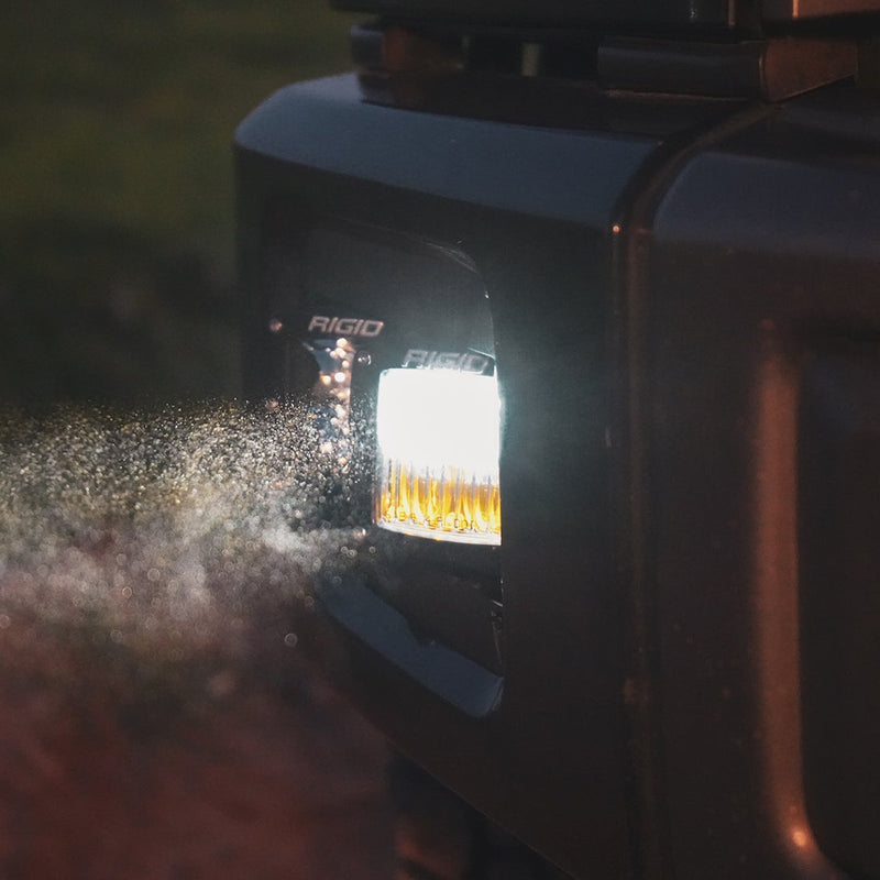 Rigid D-Series PRO SAE Pods Pair installed on a vehicle: A Rigid SAE fog pod glows white in a dark truck bumper at night as dust drifts through its beam