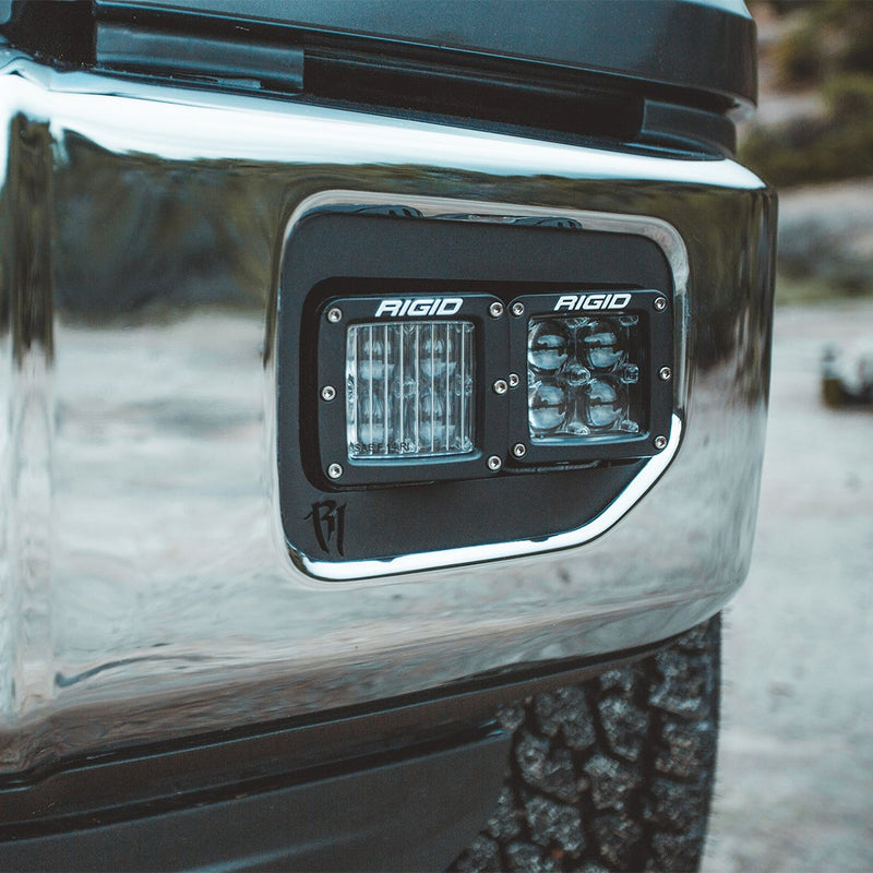 Rigid D-Series PRO SAE Pods Pair installed on a vehicle: two black Rigid pods sit side by side in the fog pocket of a chrome truck bumper, one with a fluted fog lens and one with driving optics