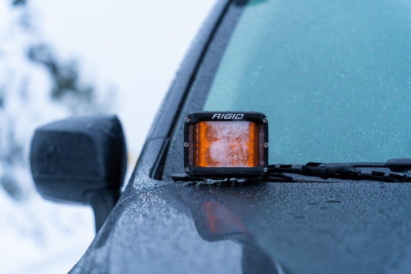 Rigid D-SS PRO Pods Pair installed on a vehicle: an amber D-SS pod glows on the cowl of a frost-covered vehicle beside its mirror
