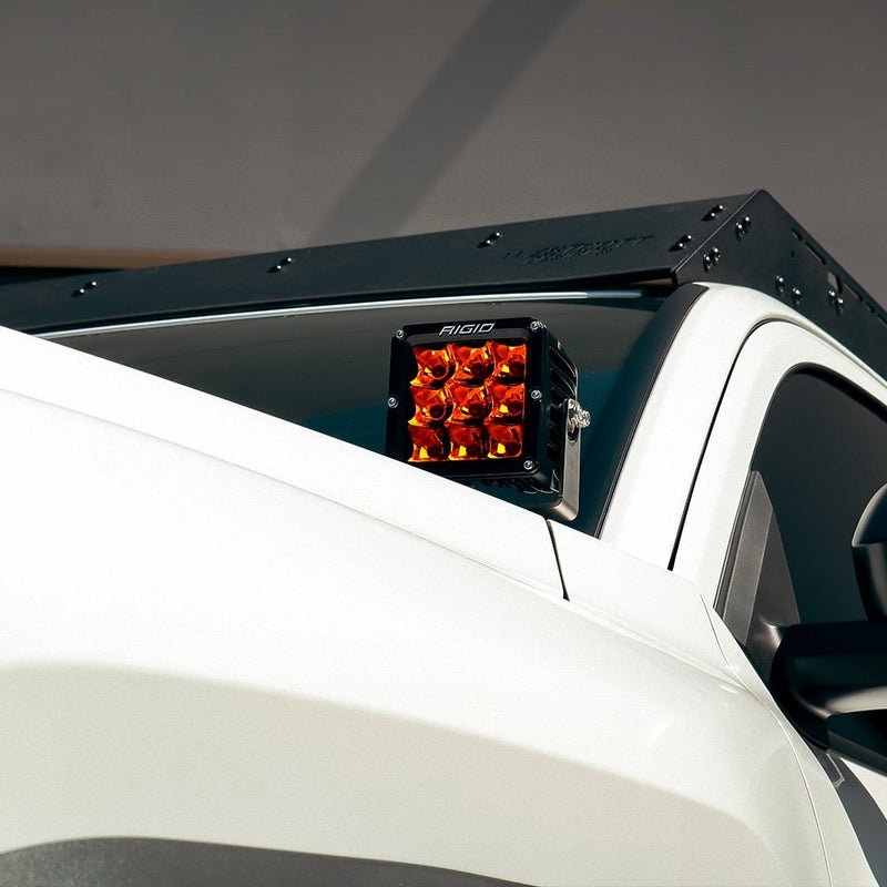 Rigid D-XL PRO Pods Pair installed on a vehicle: A D-XL pod glowing amber on an A-pillar mount below the roof rack of a white truck