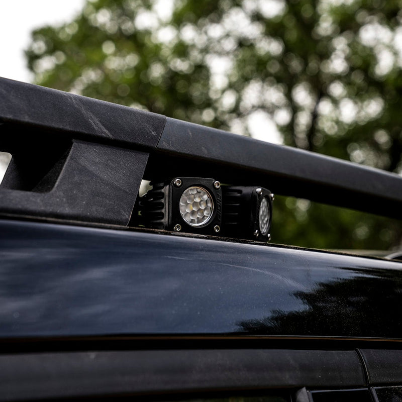 Rigid: Square black pod mounted beneath a roof rack rail on a dark vehicle, trees blurred behind