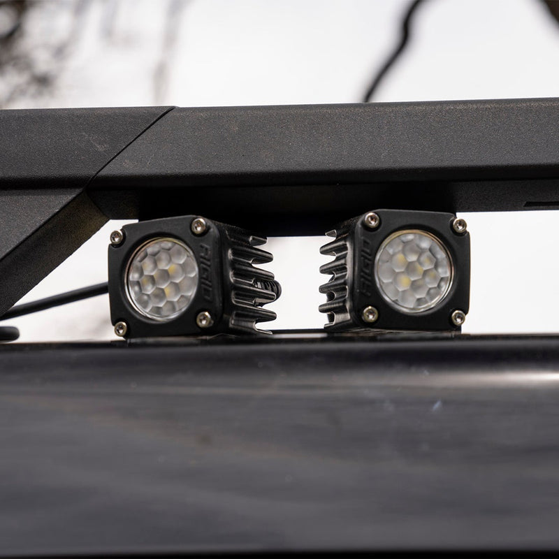 Rigid: Two square black pods mounted under a roof rack crossbar against a pale sky