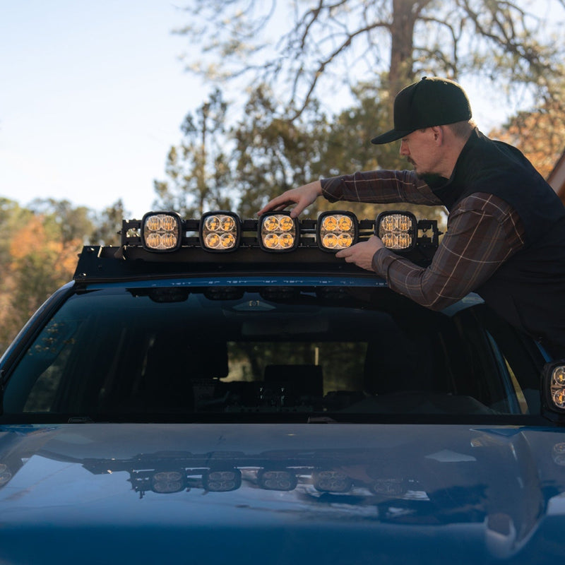 Rigid Phoenix 40" Connect Bar installed on a vehicle: A man in a cap adjusts five amber-lens pods on the roof rack of a blue vehicle