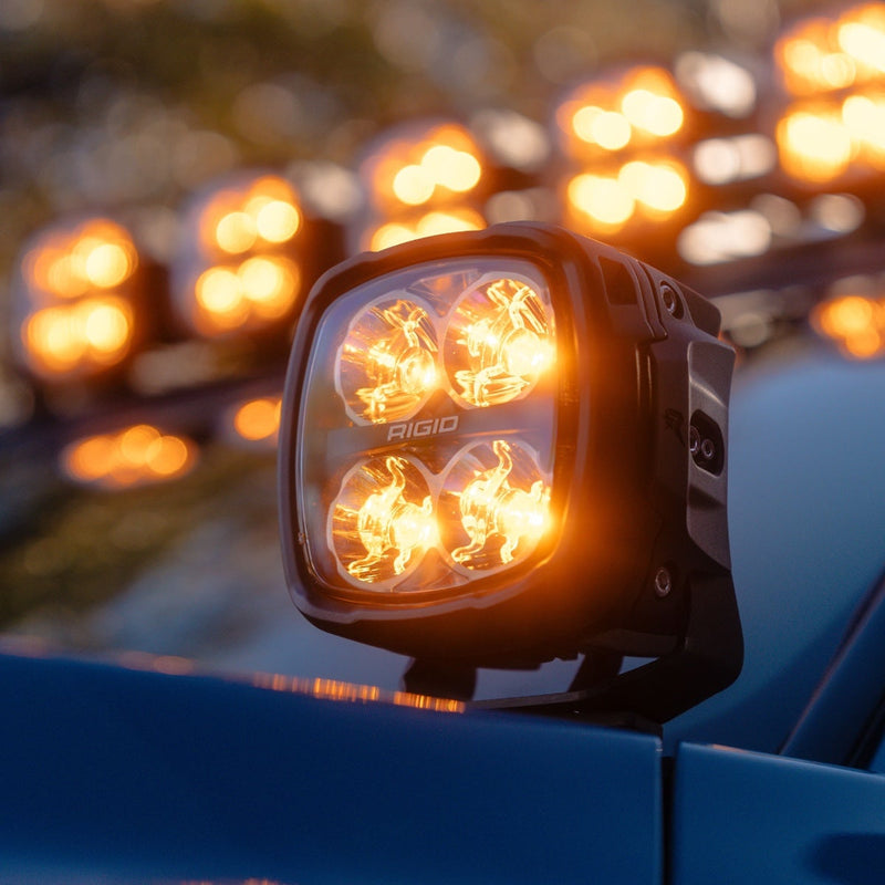Rigid Phoenix Light Pod installed on a vehicle: A pod glowing amber on a blue vehicle cowl with more lit pods blurred behind it
