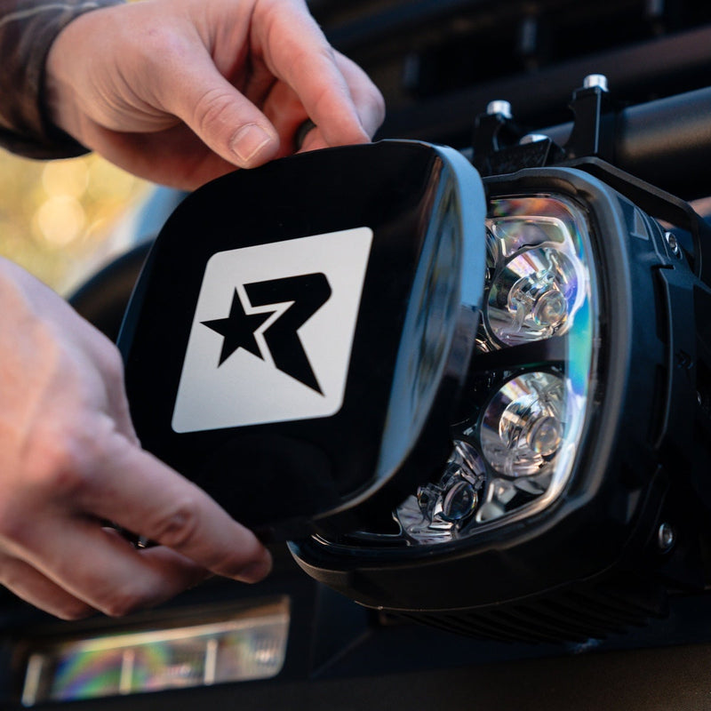 Close-up of the Rigid Phoenix XL Black Light Color: hands fit a black Rigid R cover over a large multi-optic pod mounted on a vehicle