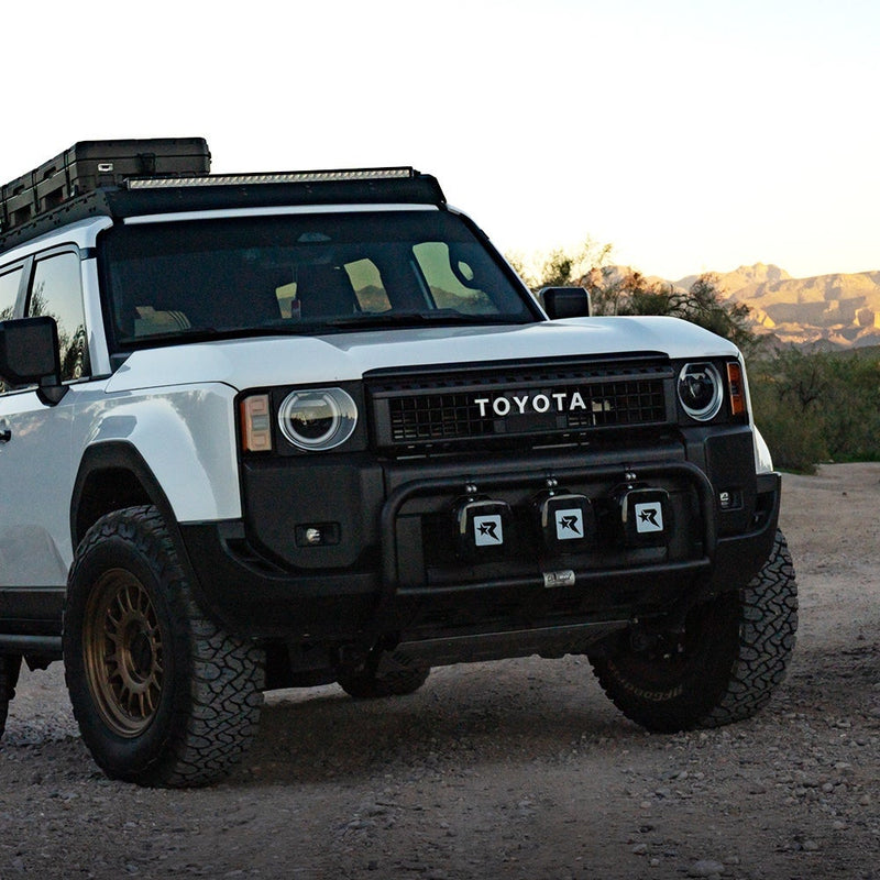 Rigid Phoenix XL Black Light Color installed on a Toyota Land Cruiser: white Toyota Land Cruiser with three black-covered pods mounted across the front bumper