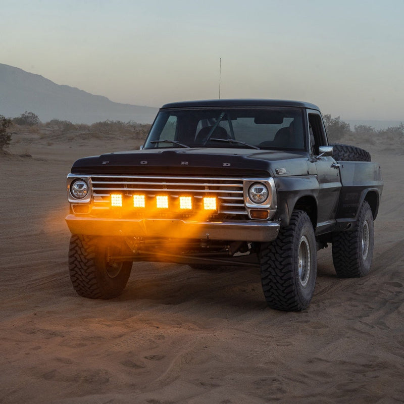 Rigid Q-Series PRO Pod installed on a Ford F-100: A classic black Ford pickup with FORD grille lettering runs five lit amber pods across its front bumper in the desert
