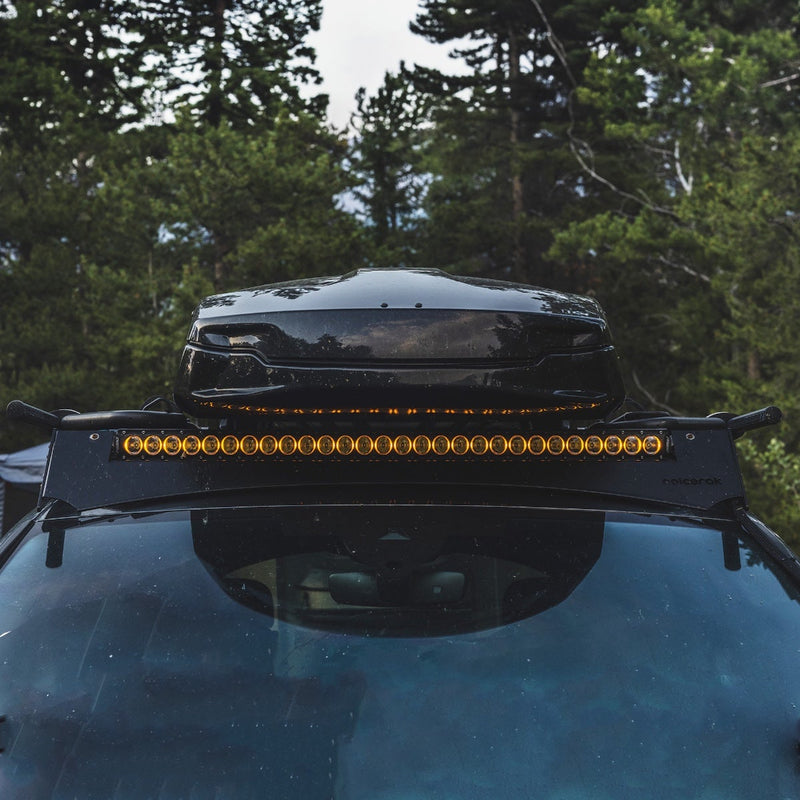 Rigid Revolve 40" Bar installed on a vehicle: A Revolve light bar with amber backlighting on a roof rack under a black cargo box, pine trees behind