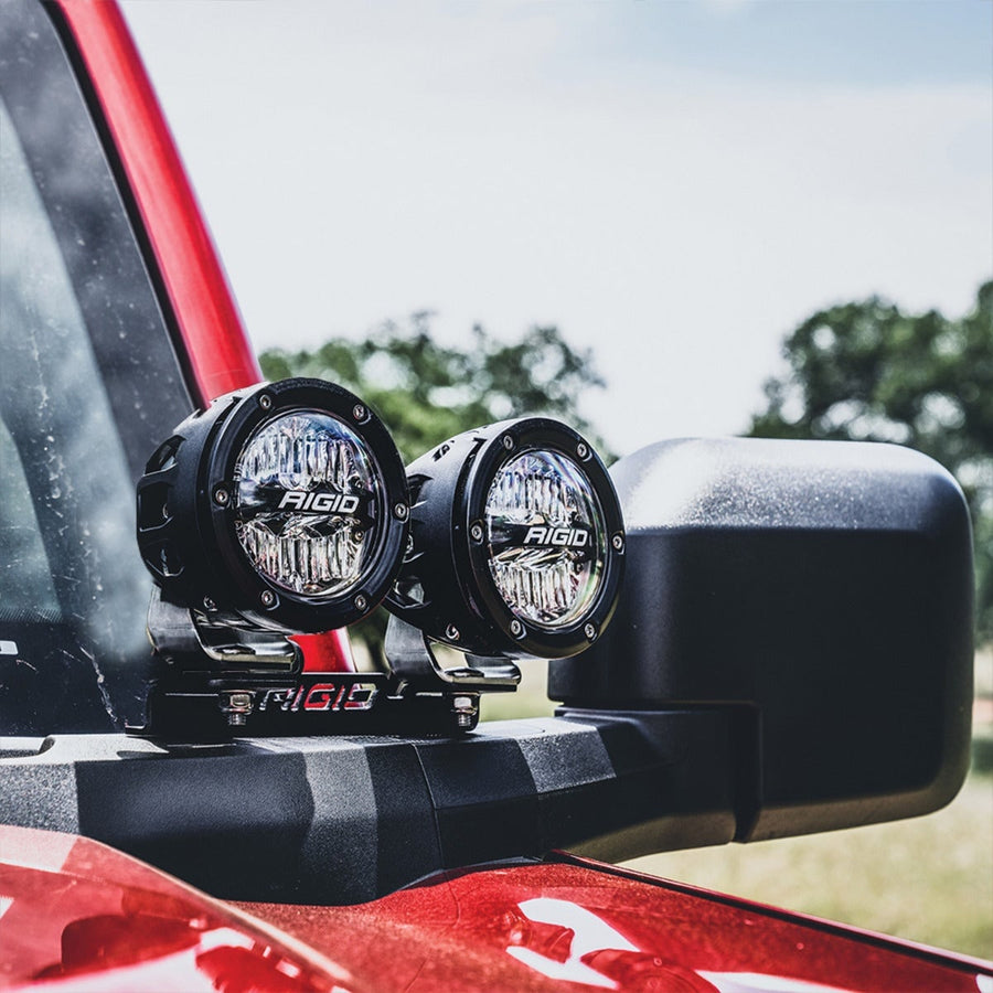 Vehicle-Specific Mounts & Brackets by Rigid: Two round black pods with clear lenses on a RIGID A-pillar mount beside the mirror of a red Ford Bronco; lights shown for illustration.