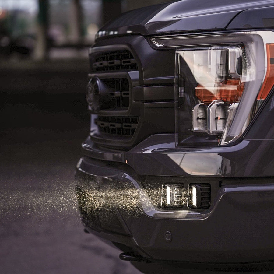 Vehicle-Specific Light Kits by Rigid: Close-up of lit square pods in the fog pocket of a dark Ford F-150 with dust in the beam.