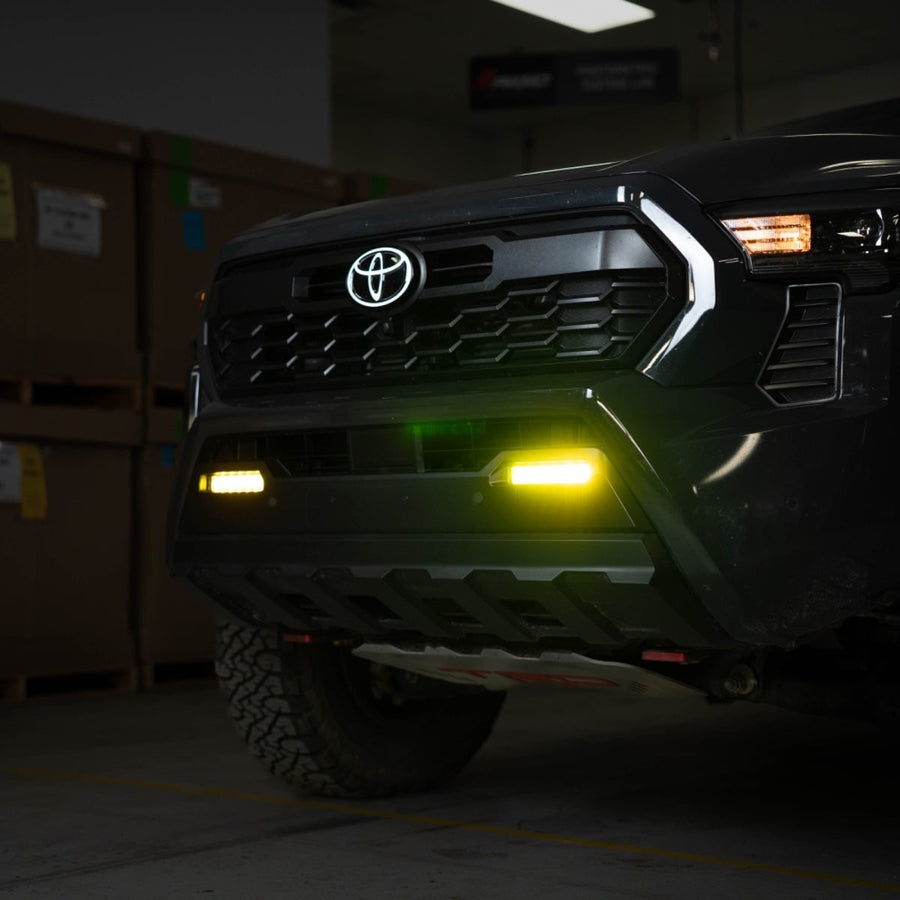 SR-Series Light Bars by Rigid: A dark Toyota pickup front in a warehouse with two selective yellow fog lights glowing in the lower bumper.