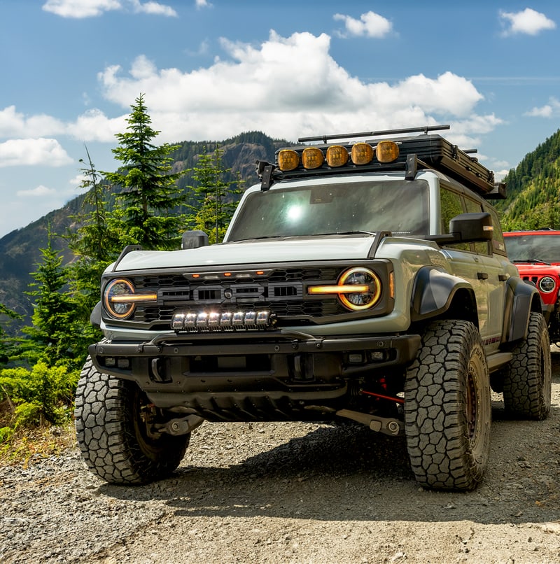 Adapt Adaptive Light Bars by Rigid: A gray Ford Bronco parked on a gravel mountain trail with a lit light bar in the front bumper and amber lights on the roof rack.