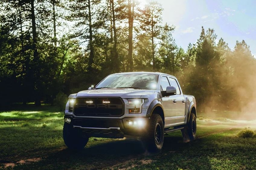 Light Mounts & Hardware by Rigid: A silver Ford Raptor pickup in a forest clearing with dust in the air and small LED lights lit in its front end.