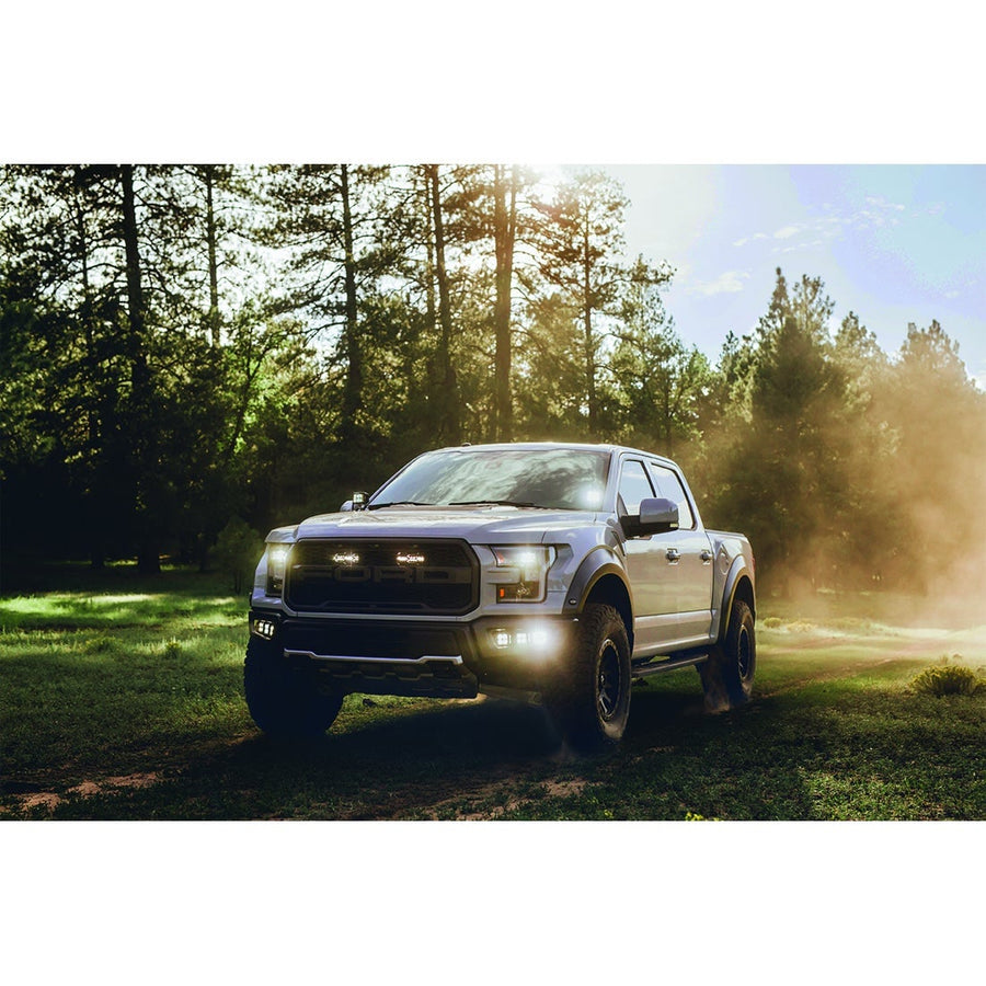 Light Mounts & Hardware by Rigid: A silver Ford Raptor pickup parked in a sunlit forest clearing with small LED lights glowing in its grille and fog pockets.