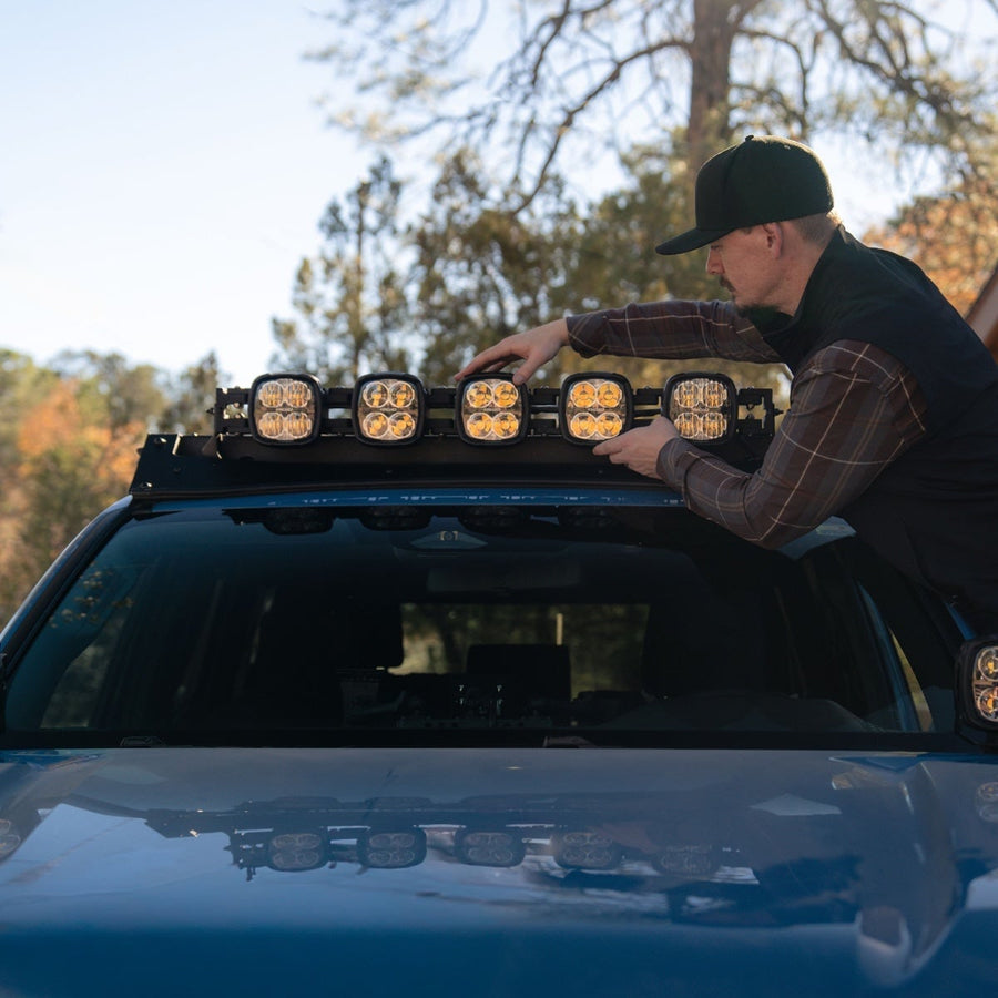 Phoenix Lights by Rigid: A man in a cap adjusts five amber-lens pods on the roof rack of a blue vehicle.