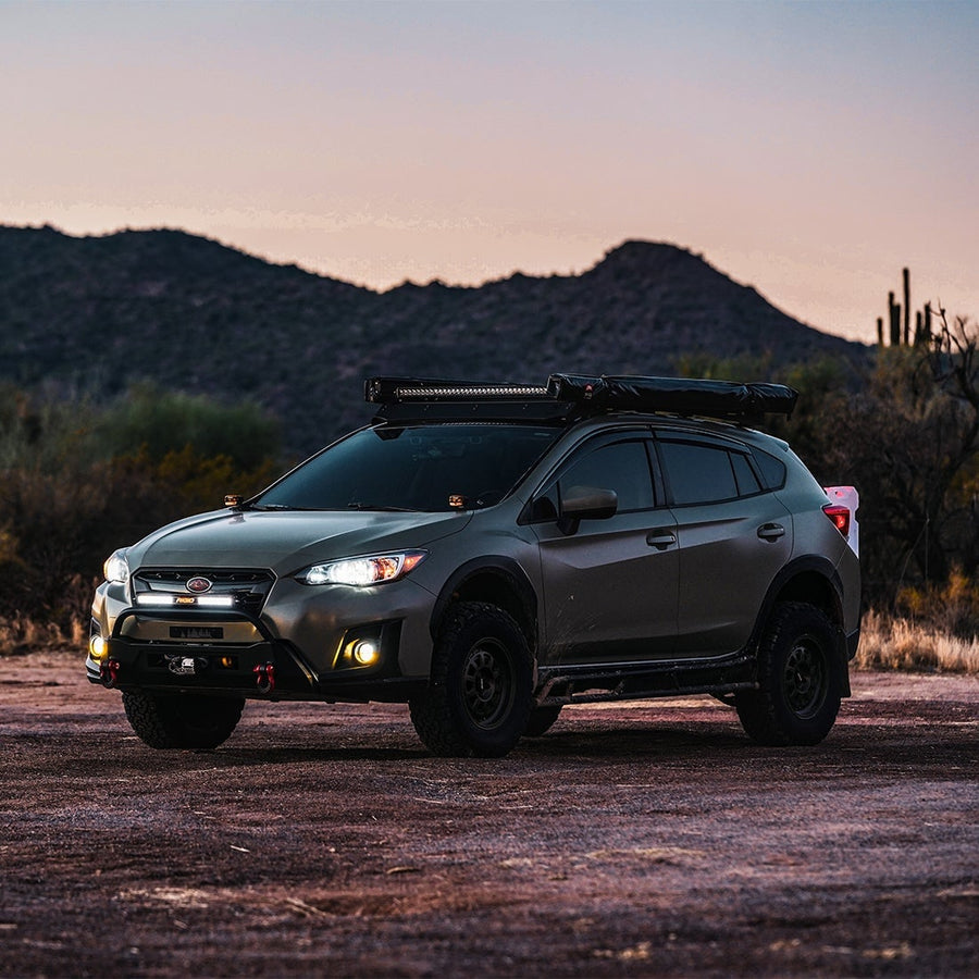 360-Series Round Lights by Rigid: A gray Subaru Crosstrek with a roof rack light bar stands on desert ground at dusk with its fog lights on.